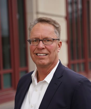 Professional senior-care advisor, Knute Rotto, wearing a navy blazer and glasses, smiling outside a red-trimmed downtown building in the Indianapolis area.