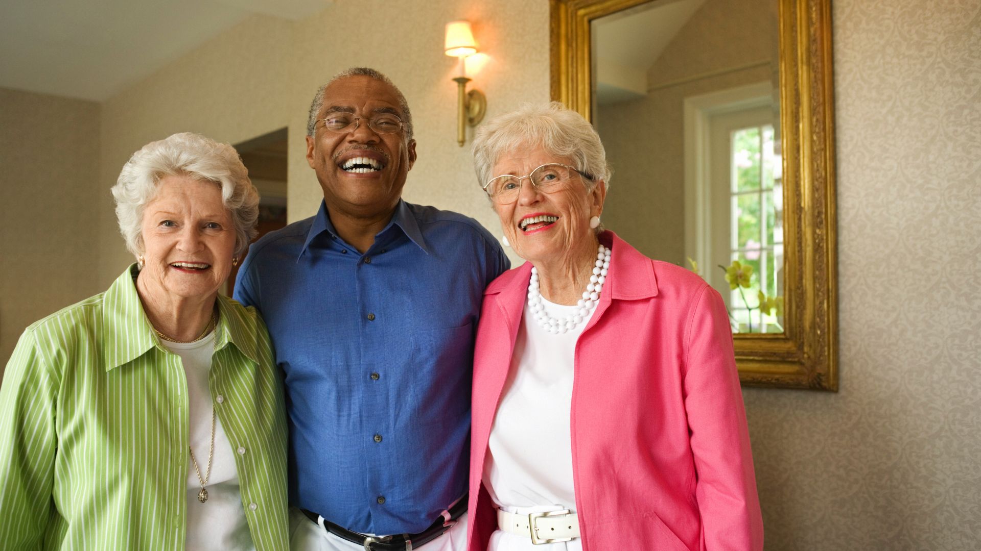 Three smiling seniors standing together indoors, dressed in bright, casual clothing, representing friendship and community in an assisted living setting.