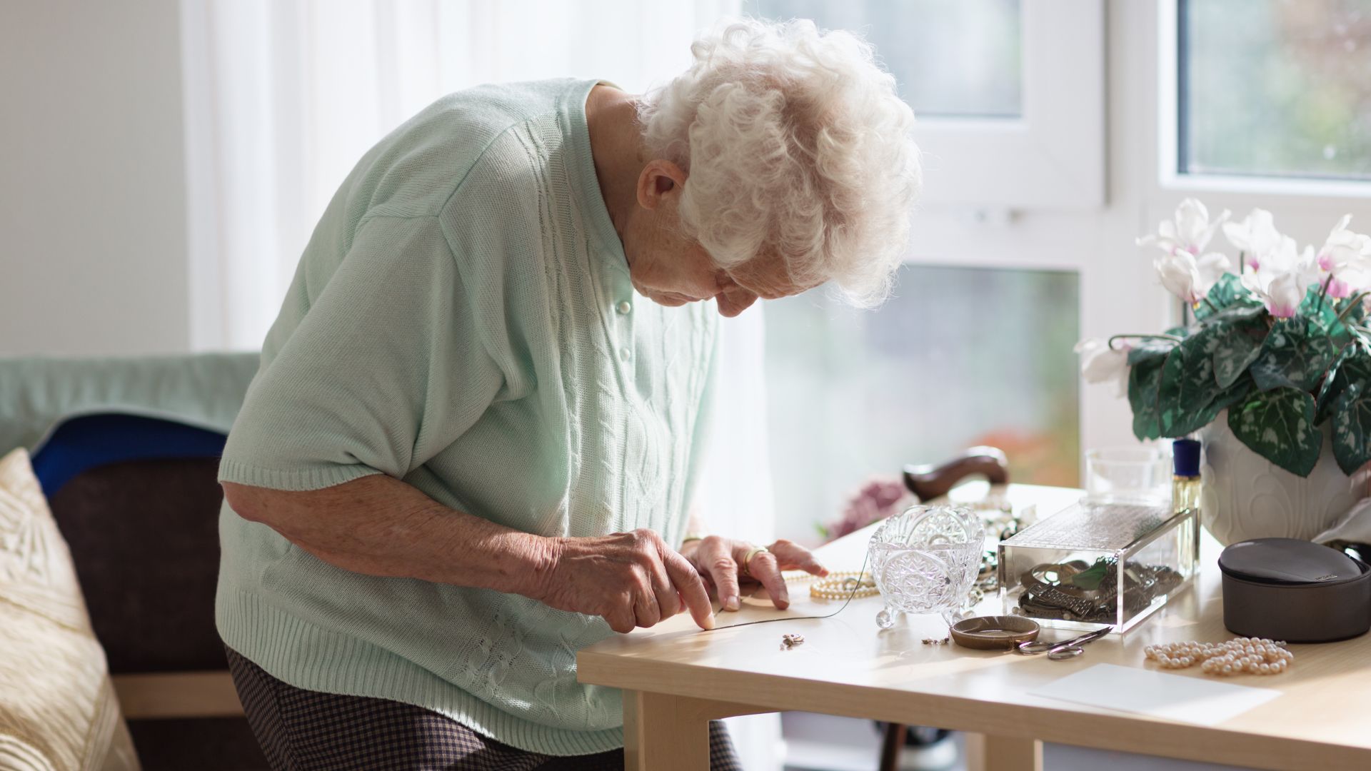 Elderly woman with white hair, wearing a light green sweater, sits at a small table working with jewelry pieces in a bright room, symbolizing memory care activities.